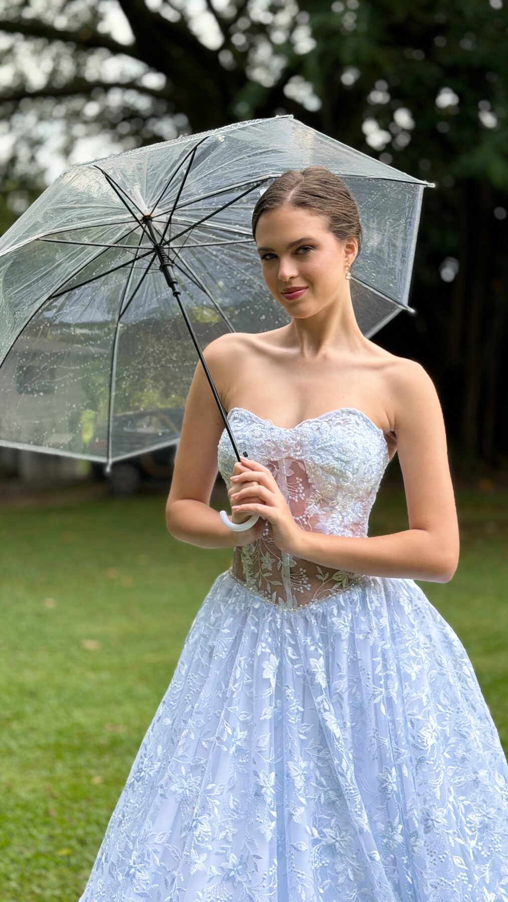 Mulher com vestido azul rendado e guarda-chuva transparente sob uma árvore