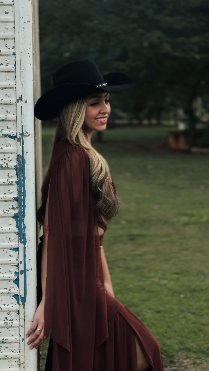 Mulher de chapéu preto e vestido marrom encostada em uma parede branca de madeira